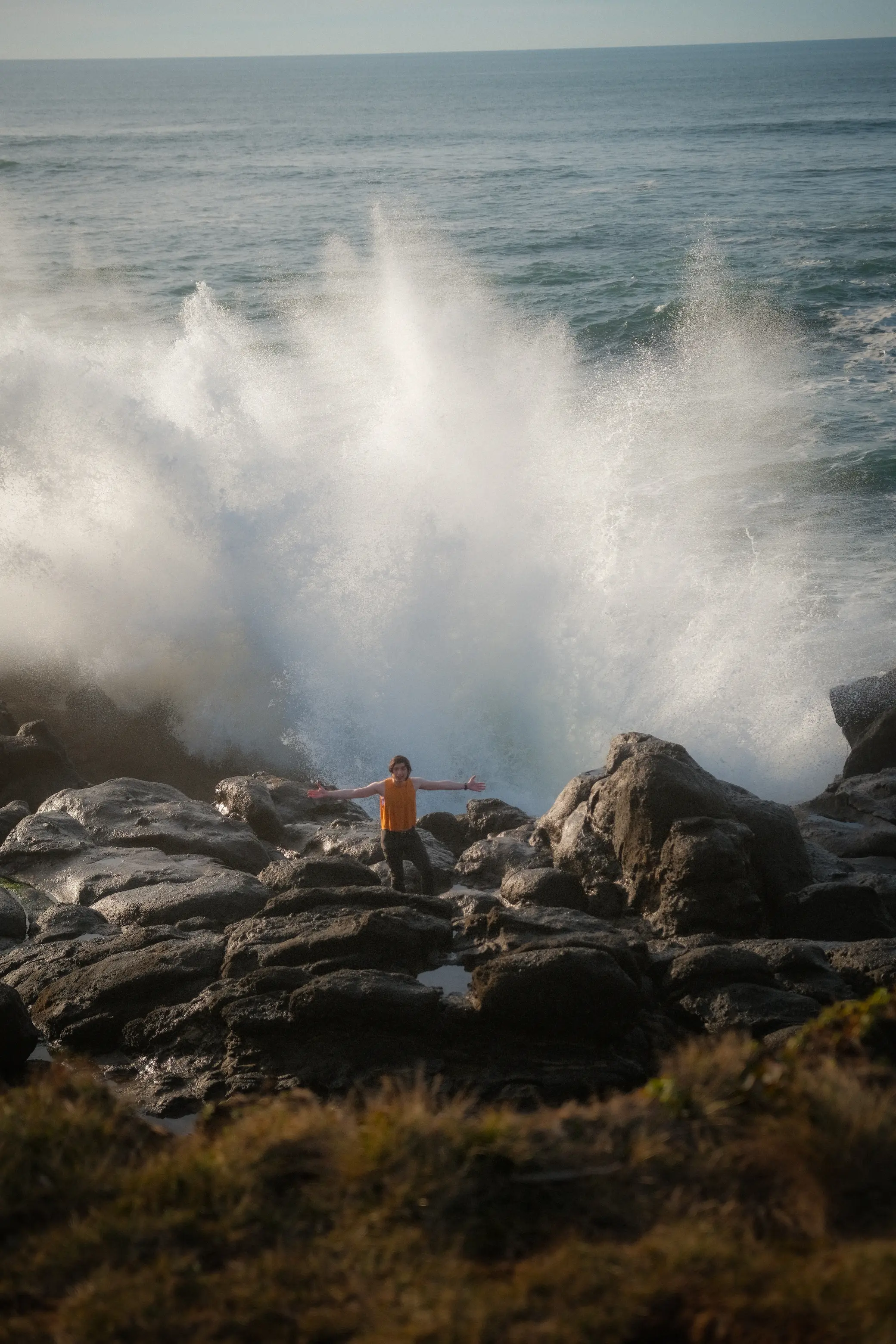 Person holding arms out while standing on rocky formation with wave splashing high behind them