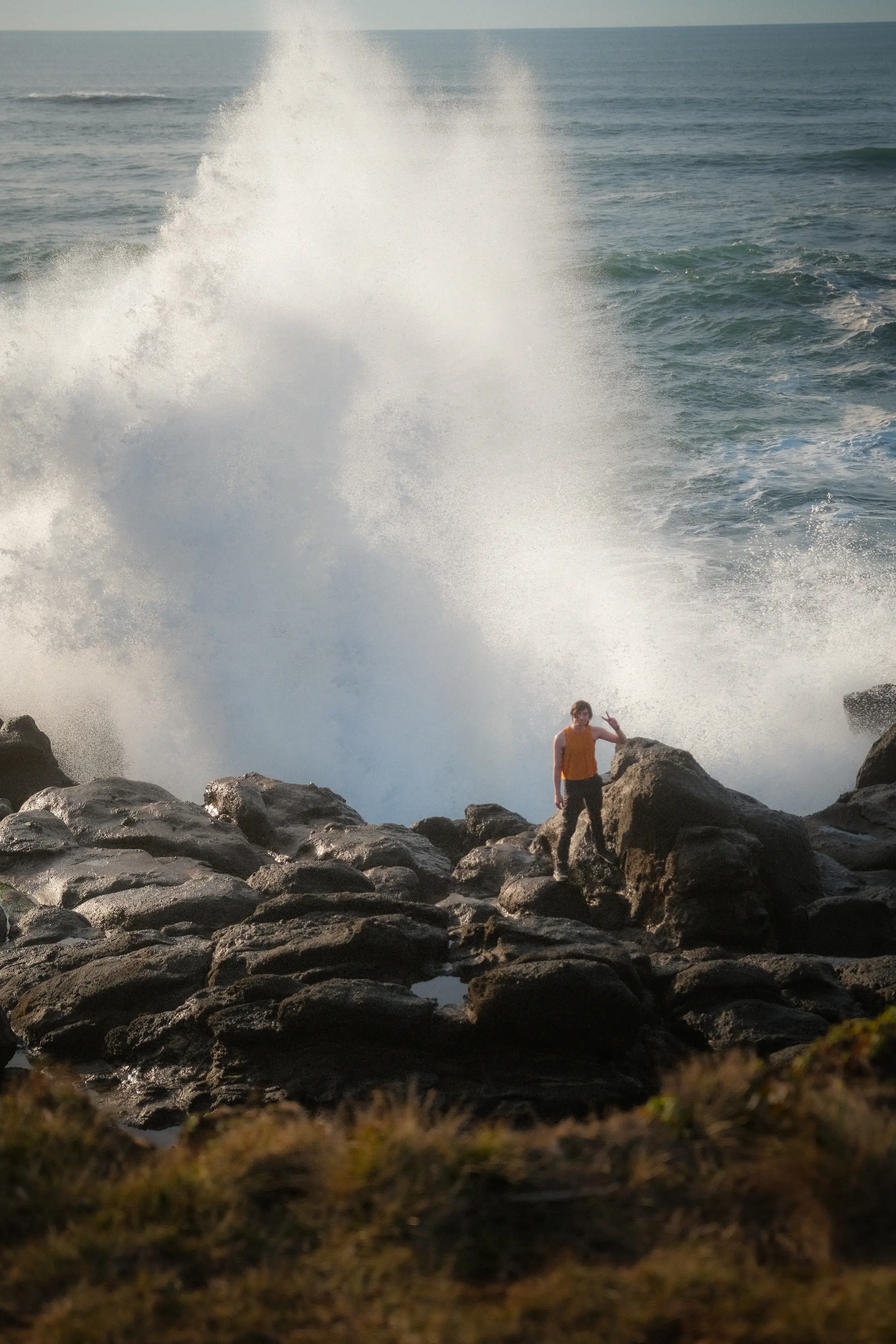Person holding peace sign on left hand while standing on rocky formation with wave splashing high behind them