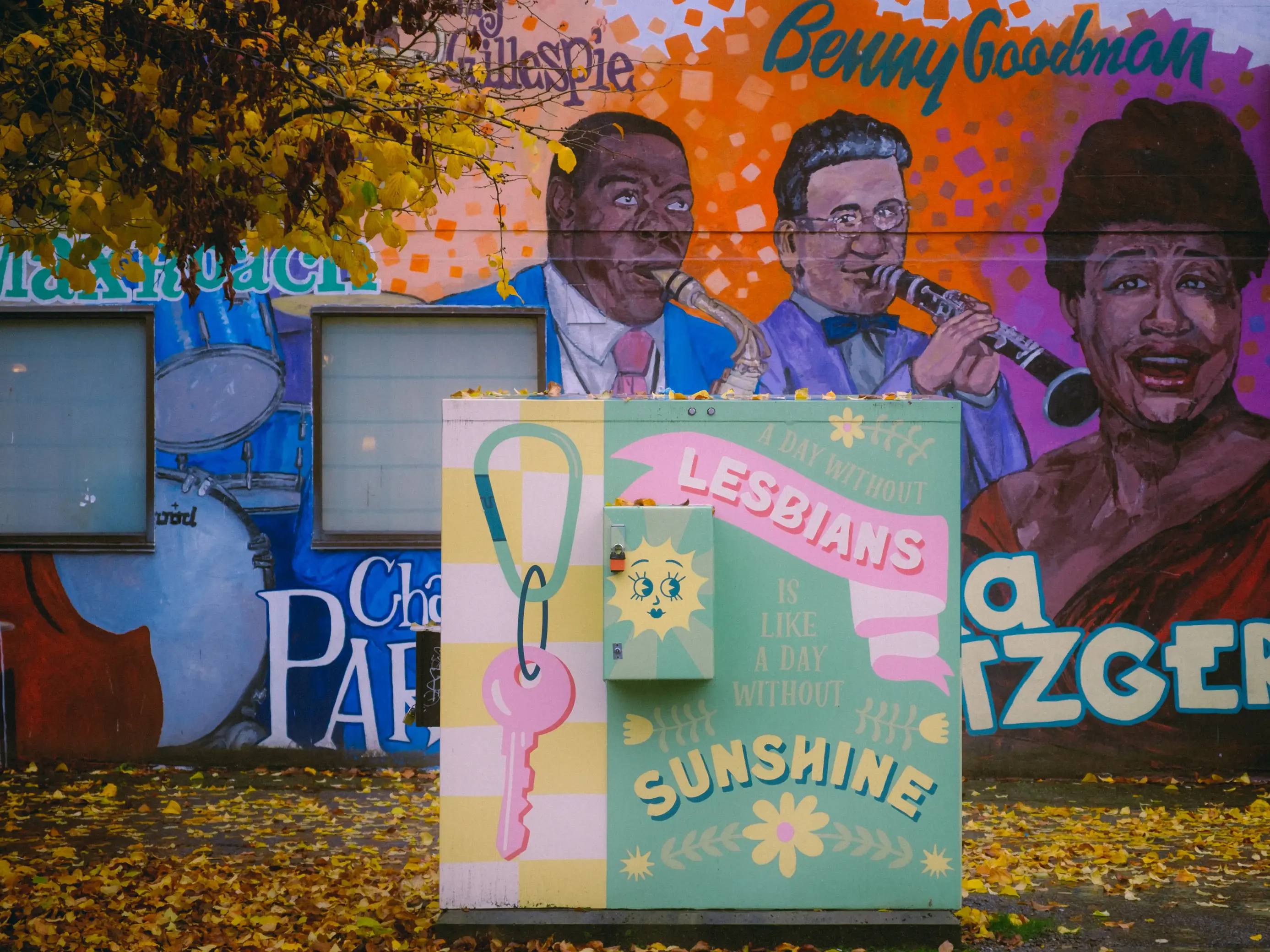 photo of a mural that says a day without lesbians is like a day without sunshine. there is a carabiner attached to a key behind that mural, there is another mural of famous jazz musicians there are yellow leaves on the ground