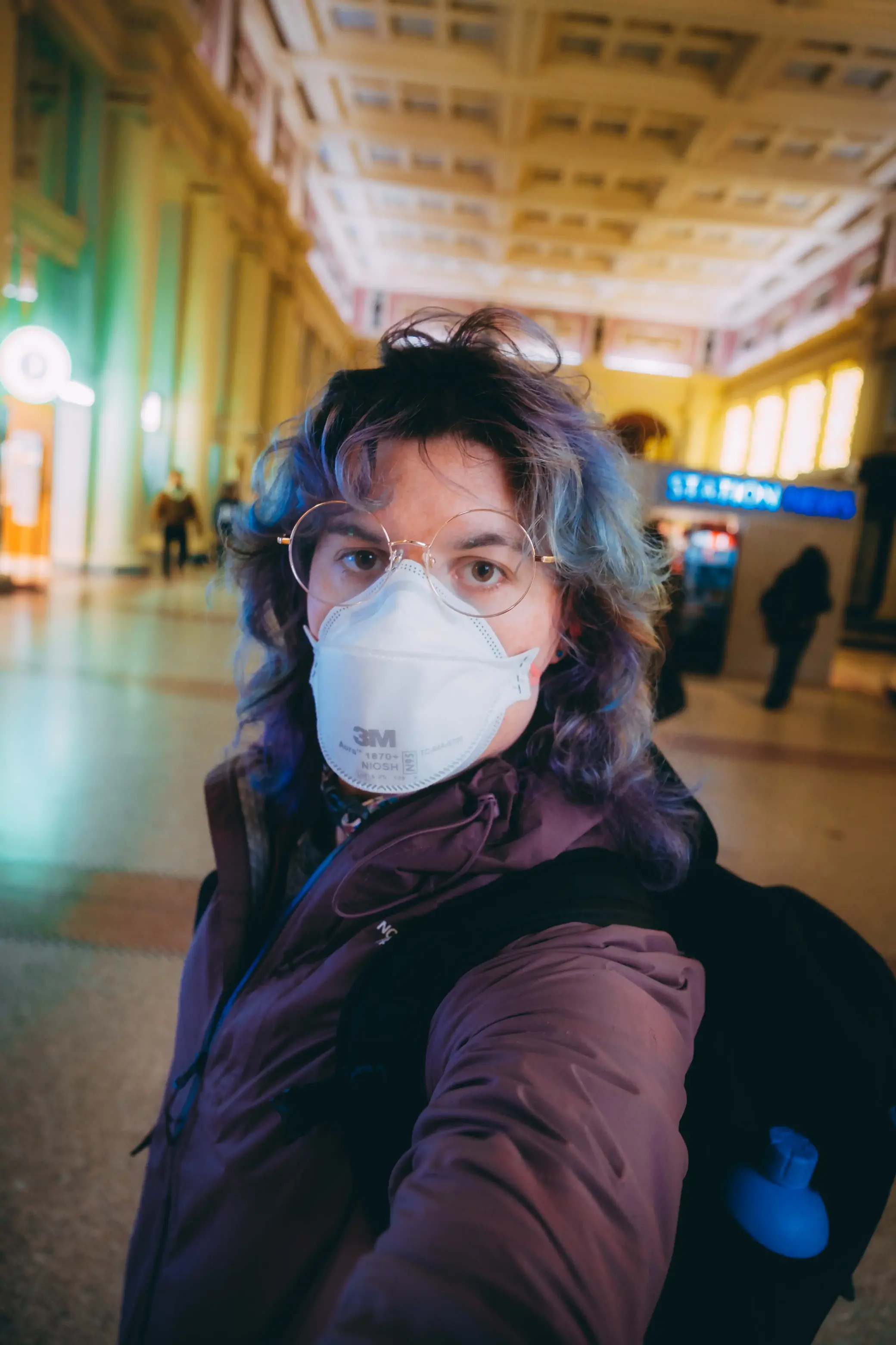 the author taking a selfie in Waterfront station in Vancouver BC she's wearing a purple rain jacket, has fading purple hair that shows some blue, is wearing a backpack, glasses, and a 3M N95 mask